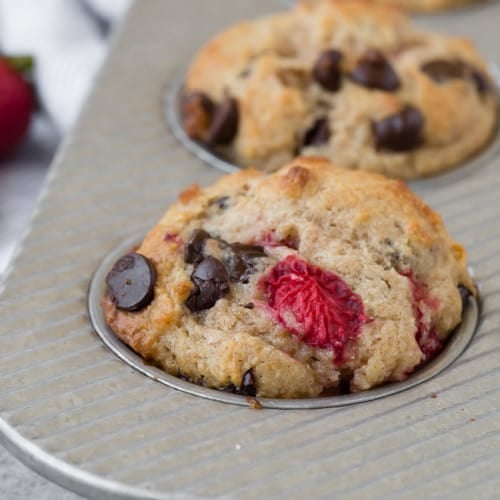 Close-up image of a muffin flecked with fresh strawberries and gooey dark chocolate chips. More strawberries and chocolate chips are pictured in the background.