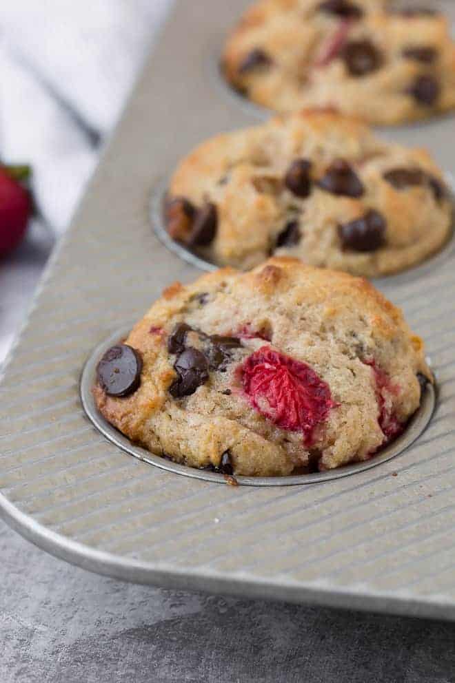 Close-up image of a muffin flecked with fresh strawberries and gooey dark chocolate chips. More strawberries and chocolate chips are pictured in the background.