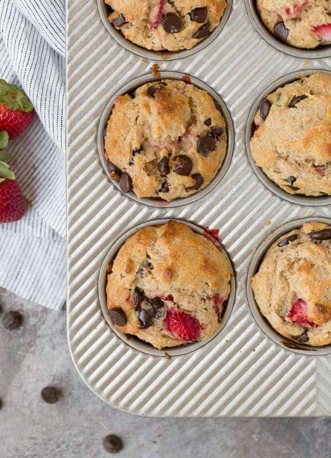  Overhead-Bild von Muffins in einer Dose. Sie sind mit Schokoladenstückchen und Erdbeeren gesprenkelt, und es gibt zusätzliche Schokoladenstückchen und Erdbeeren an der Seite. 