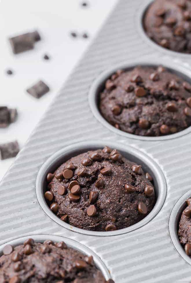 Close up view of chocolate muffins in a muffin pan. Chocolate chunks and chocolate chips are pictured in the background, on a white backdrop.