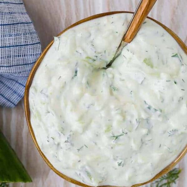 Overhead view of creamy yogurt sauce in a bowl on a wooden background. Cucumbers and dill are placed around the bowl.