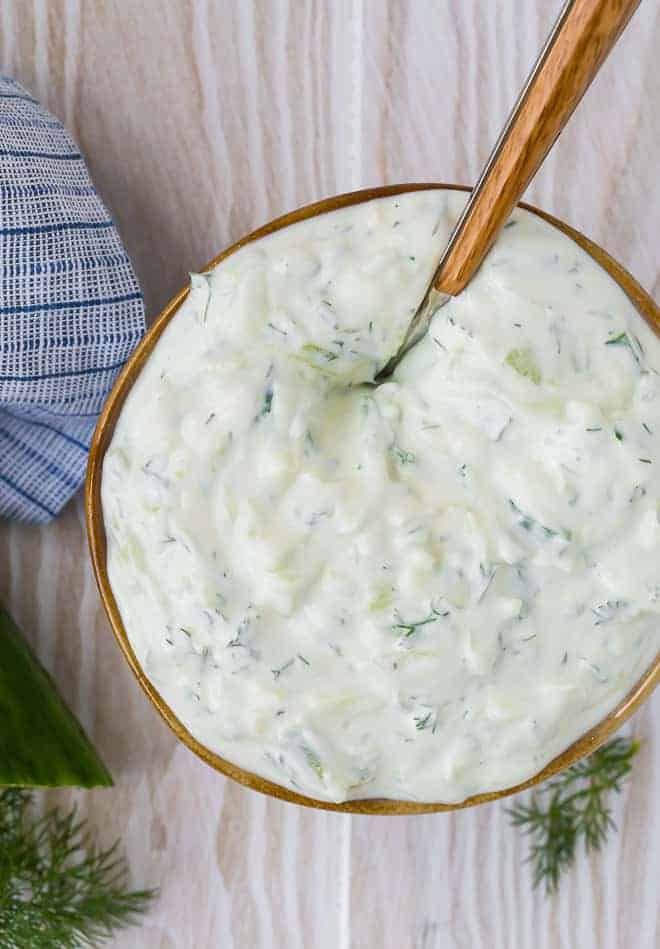 Overhead view of creamy yogurt sauce in a bowl on a wooden background. Cucumbers and dill are placed around the bowl.