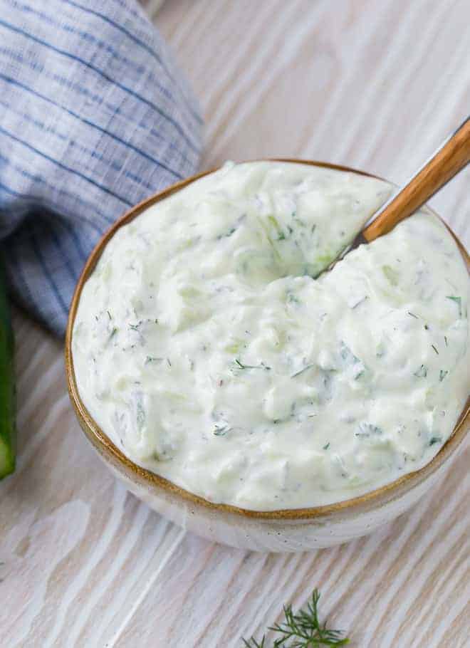 A creamy yogurt sauce made with cucumbers, dill and lemon in a natural colored bowl with a wooden spoon inserted. 