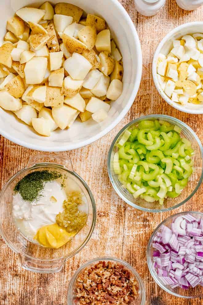 Overhead view of bowls of ingredients, clockwise from top, cooked and diced potatoes, chopped boiled eggs, chopped celery, chopped red onion, crumbled bacon, and the last bowl is filled with a few ingredients: mayo, mustard, relish, dill.