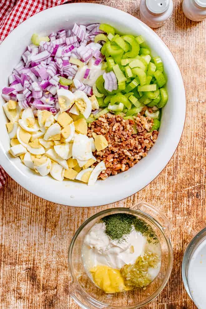 Overhead view of a large bowl filled with chopped boiled eggs, chopped red onion, chopped celery, crumbled bacon. Another small glass bowl sits next to it, filled with a mayonnaise based dressing.