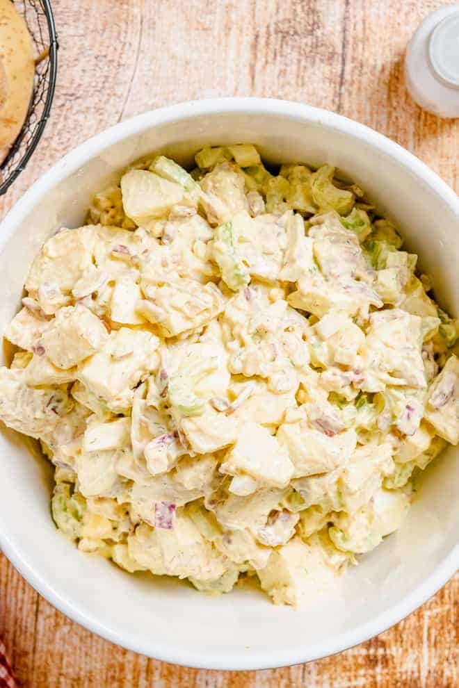 Overhead view of a large white bowl on a wooden background. The bowl is filled with a creamy salad made with potatoes, eggs, celery, bacon, and more.