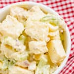 Close up overhead view of a bowl of diced potatoes, celery, onion, bacon, and boiled eggs tossed in a creamy light yellow dressing with flecks of green.