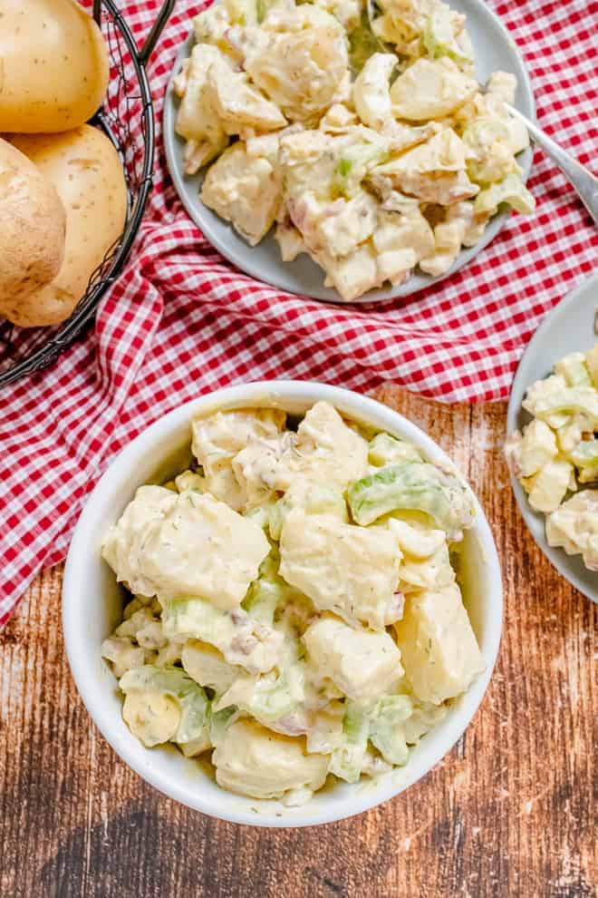 Overhead view of creamy potato salad in two plates and one bowl, with whole unpeeled potatoes in the background of the image.