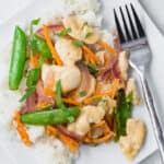 Overhead view of a white plate filled with white rice, chicken, sugar snap peas, carrots, and red onions in stir fry form. It is garnished with fresh basil ribbons.