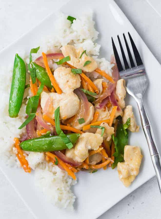 Overhead view of a white plate filled with white rice, chicken, sugar snap peas, carrots, and red onions in stir fry form. It is garnished with fresh basil ribbons.