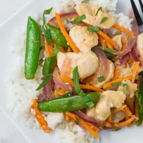 Overhead view of a white plate filled with white rice, chicken, sugar snap peas, carrots, and red onions in stir fry form. It is garnished with fresh basil ribbons.