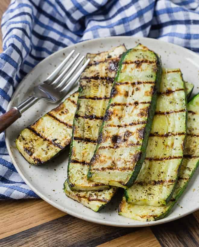 An earth-tone plate on a wooden background with a blue and white linen. The plate is full with grilled zucchini slices. 