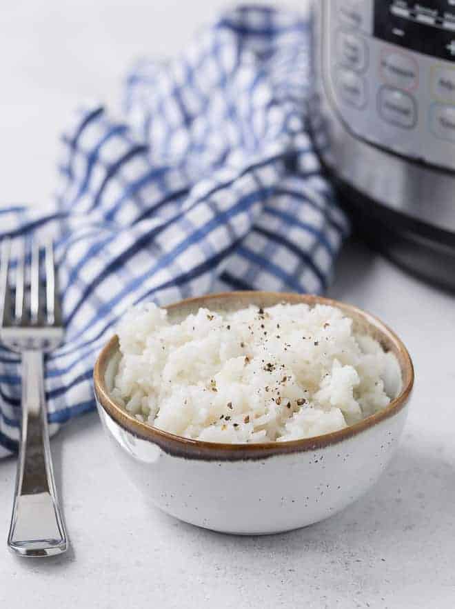 Cooked white rice in a bowl with a fork next to it, a blue and white linen and an instant pot are in the background.