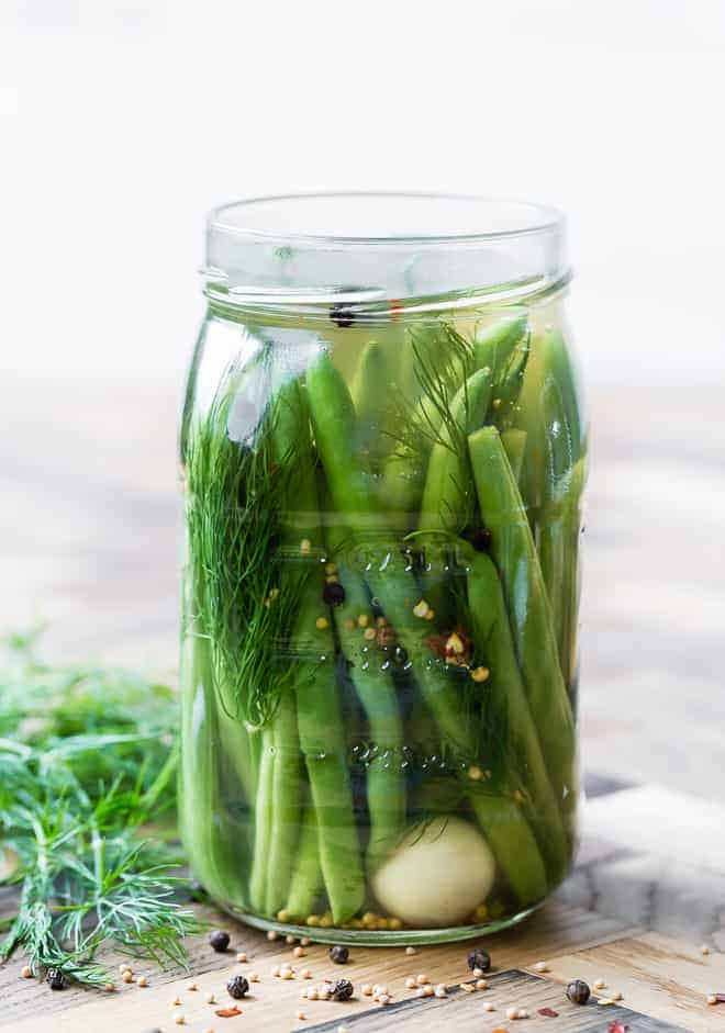 Large glass jar of pickled green beans on a wooden background with ingredients scattered around.