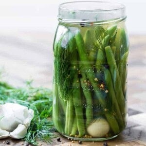 Large glass jar of green beans, dill, a clove of garlic, spices, and pickling liquid. Additional dill and garlic are pictured in the background on a wooden surface.
