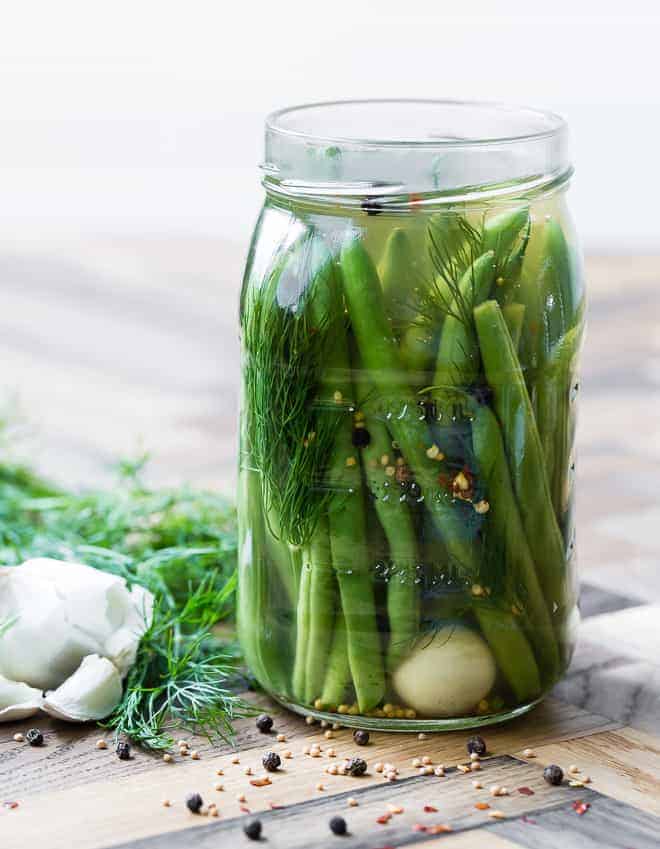 Large glass jar of green beans, dill, a clove of garlic, spices, and pickling liquid. Additional dill and garlic are pictured in the background on a wooden surface.