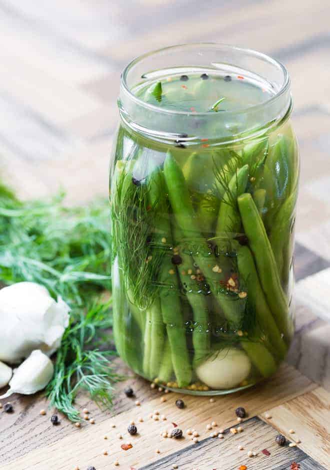A glass jar filled with green beans and pickling ingredients on a wooden background, with ingredients such as red pepper flakes, peppercorns, mustard seeds, garlic and dill also visible.