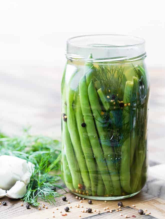 Large glass jar full of pickled green beans on a wooden surface. Dill, garlic, and spices are also pictured.