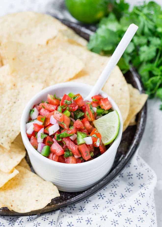 A small white bowl surrounded by tortilla chips. The bowl is filled with chopped tomatoes, onions, cilantro, and jalapeno.