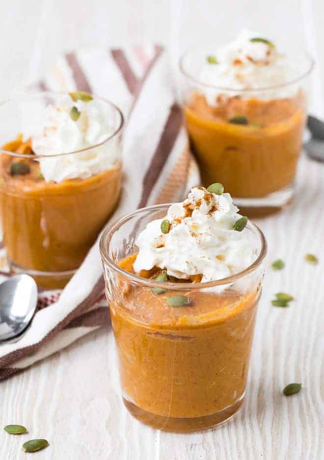 Three small glass dishes filled with orange pumpkin pudding, topped with whipped cream and garnished with pepitas and cinnamon.