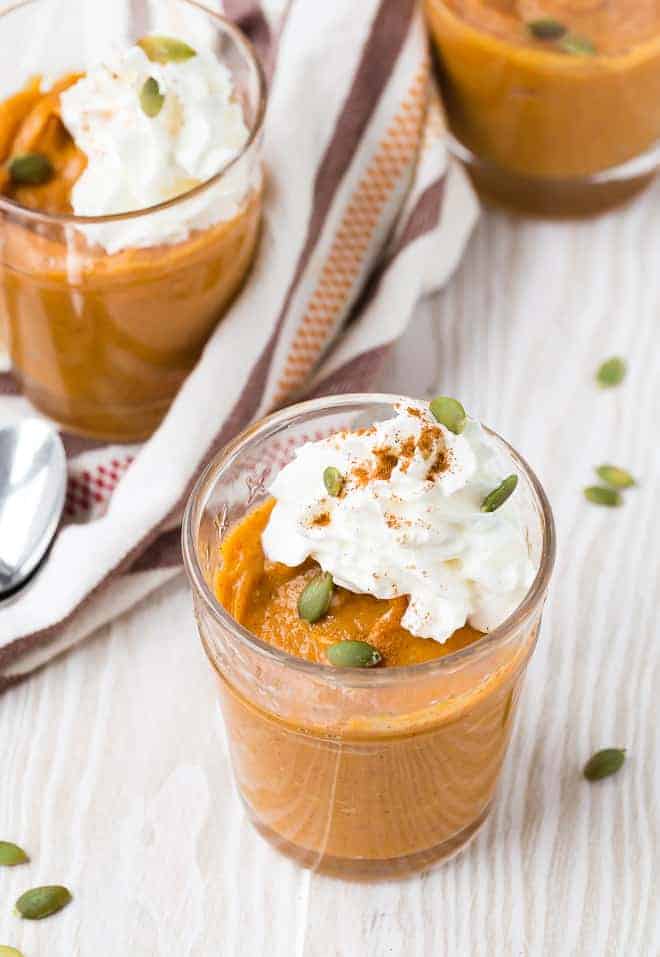 Three small glass dishes filled with orange pumpkin pudding, topped with whipped cream and garnished with pepitas and cinnamon.