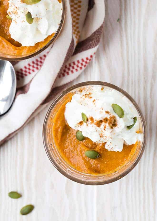 Overhead view of a small clear glass filled with orange pumpkin pudding, topped with whipped cream and garnished with pepitas and cinnamon.