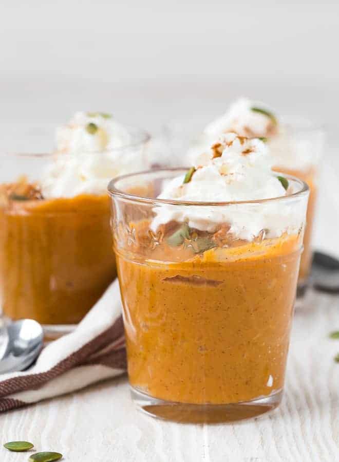 Three small glass dishes filled with orange pumpkin pudding, topped with whipped cream and garnished with pepitas and cinnamon.