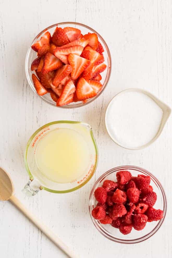 Overhead image of four glass containers, one with strawberries, one with sugar, one with lemon juice, and one with fresh raspberries.