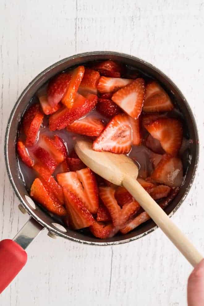 Overhead view of a sauce pan full of strawberries and red liquid, being stirred with a wooden spoon.