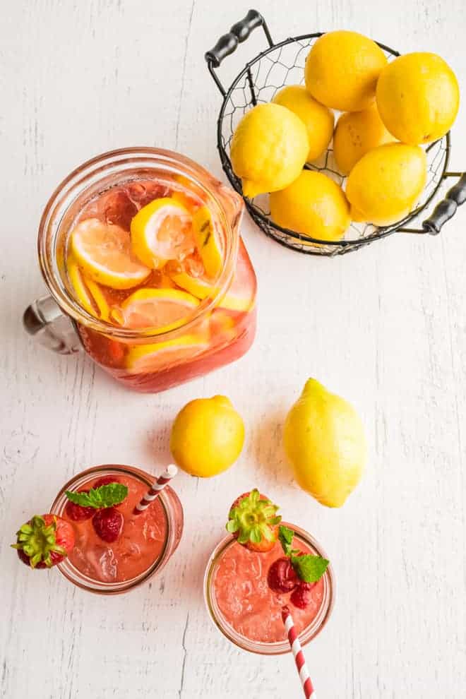 Overhead view of two glasses of pink lemonade, a pitcher, a bowl of lemons, and two lemons on a white wooden background.