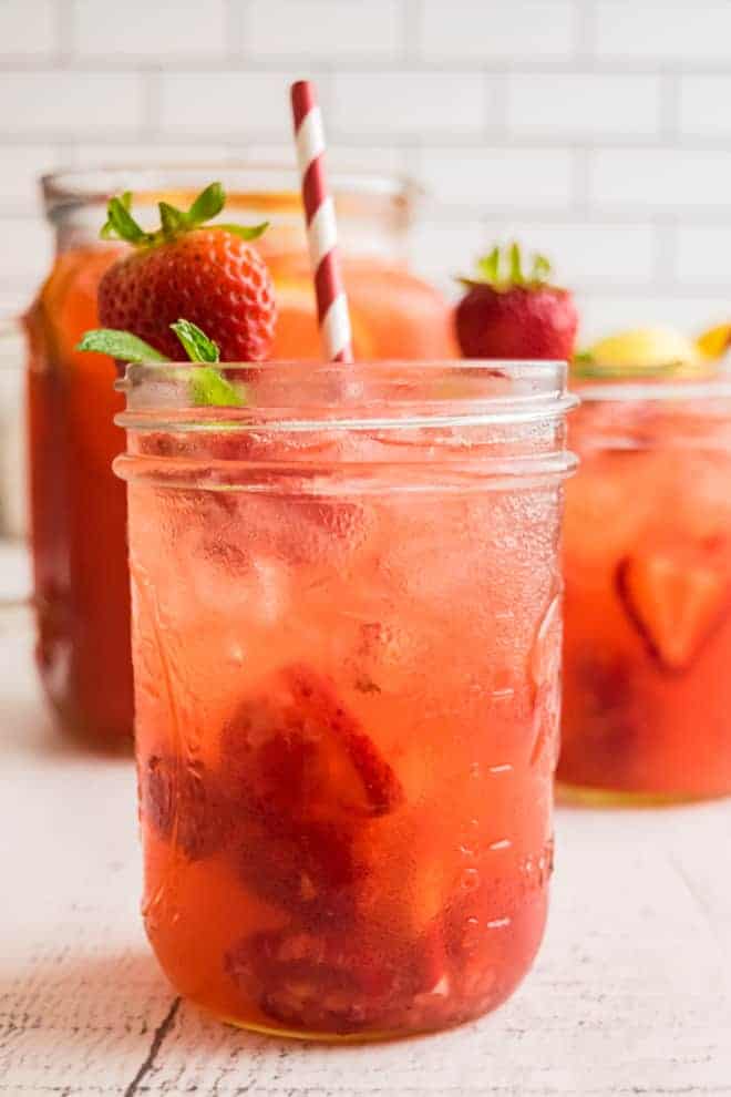 A light red drink on ice in a small mason jar. It is garnished with a fresh strawberry, mint leaves, and a red and white striped paper straw.