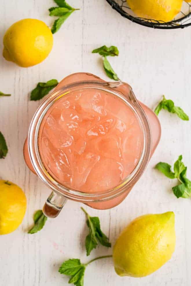 Overhead view of pink lemonade in a pitcher. Mint sprigs and lemons are scattered around.