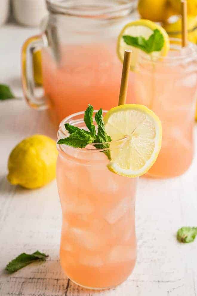 A tall glass of watermelon lemonade with a straw, garnished with lemon slices and fresh mint. Ingredients are pictured in the background. 