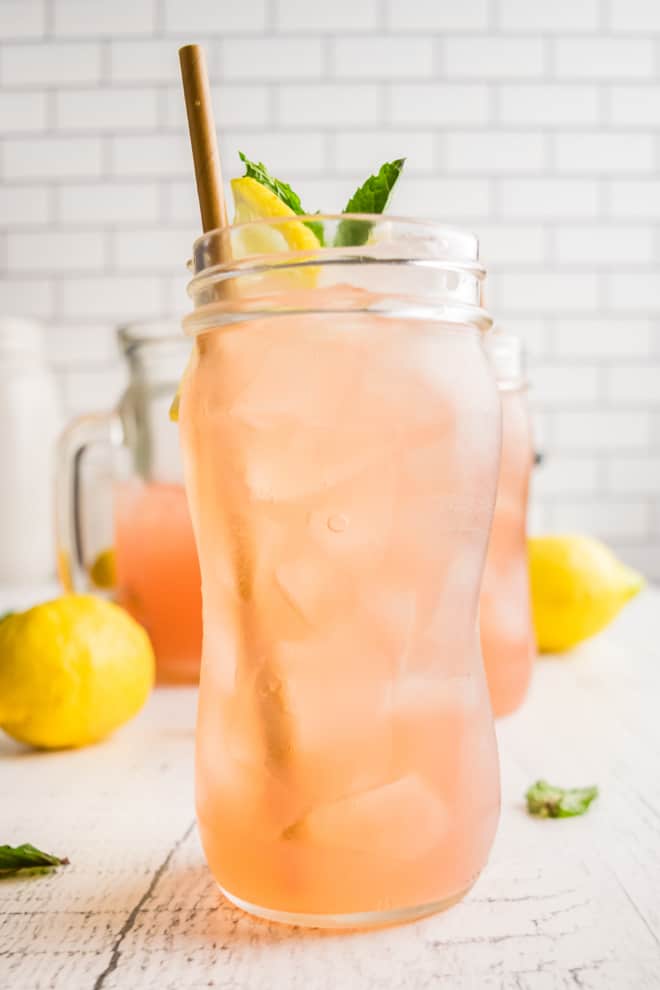 A tall glass of watermelon lemonade with a straw, garnished with lemon slices and fresh mint. Ingredients are pictured in the background. 