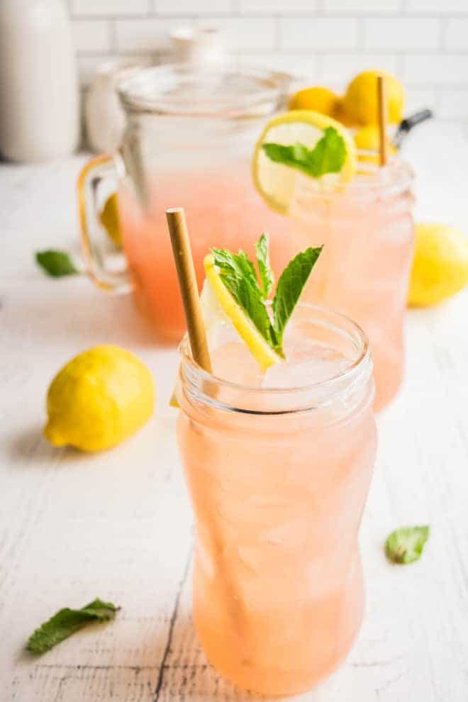 A tall glass of watermelon lemonade with a straw, garnished with lemon slices and fresh mint. Ingredients are pictured in the background. 