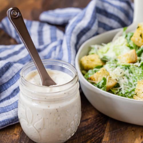 Small jar of caesar salad dressing in front of a bowl of caesar salad on a wooden background.