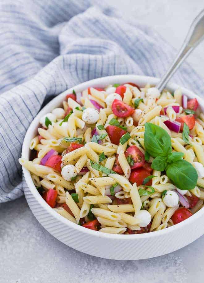 A white bowl full of mini penne pasta, grape tomatoes, pearl mozzarella, and fresh basil. A large silver serving spoon is also in the bowl.