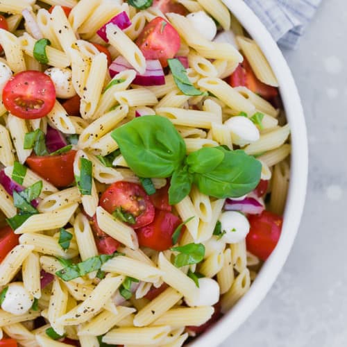 Close up partial view of a bowl of pasta salad with tomatoes, fresh mozzarella, red onion, and fresh basil.