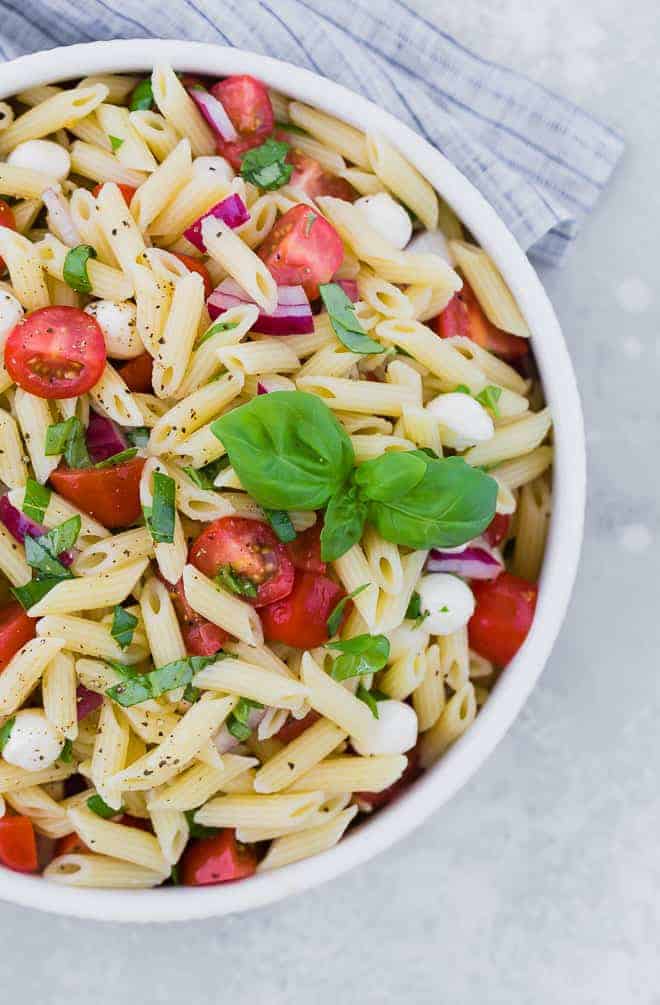 Close up partial view of a bowl of pasta salad with tomatoes, fresh mozzarella, red onion, and fresh basil.