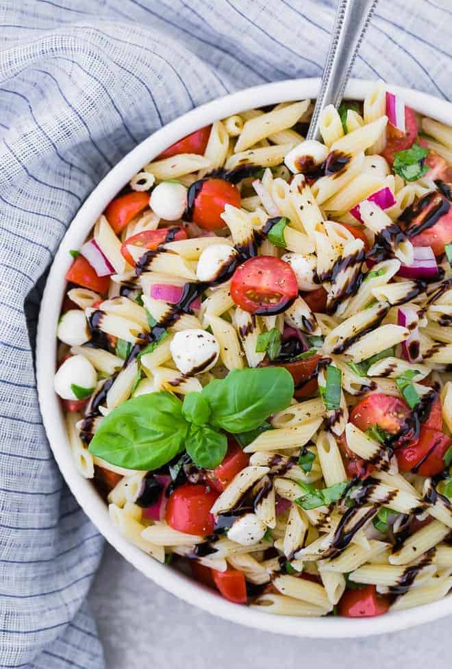 A large white bowl full of caprese pasta salad with tomatoes, mozzarella, red onions, and fresh basil. The salad is drizzled with balsamic reduction.