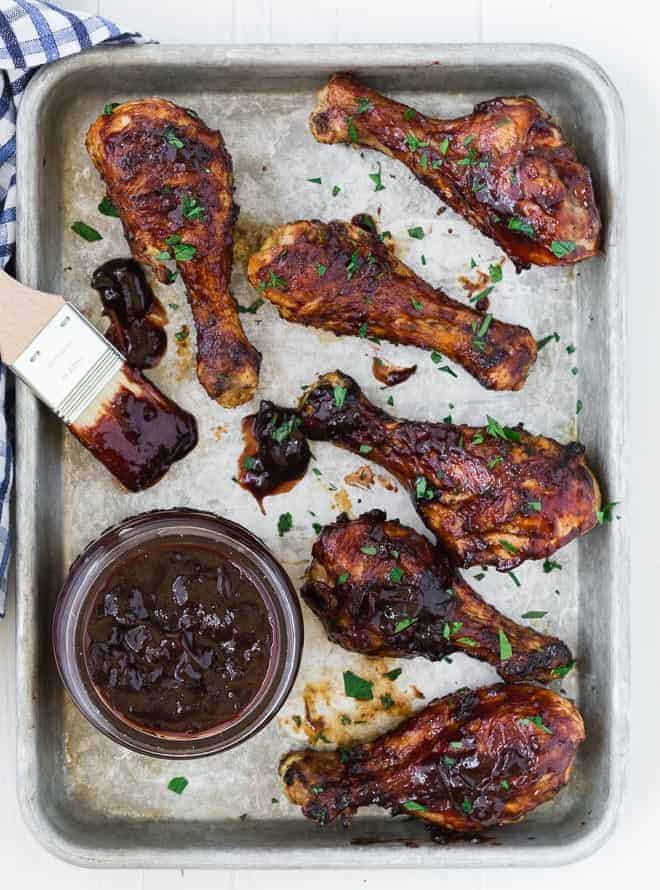 Overhead view of chicken drumsticks on a metal sheet pan with a jar of homemade bbq sauce and a brush also pictured.