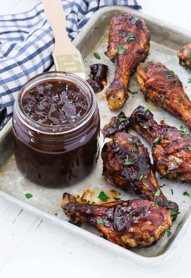 A sheet pan with grilled chicken drumsticks, and a jar of homemade barbecue sauce. A blue and white checkered linen is also pictured.