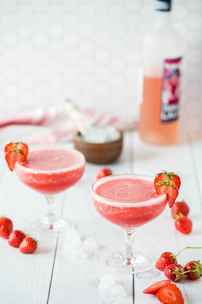 Two glasses of frose, garnished with strawberries. Fresh strawberries, ice cubes, and a bottle of wine are also pictured.