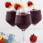 Three long stem wine glasses filled with frozen sangria, and topped with a fresh strawberry and orange slice. A paper straw is in each glass.