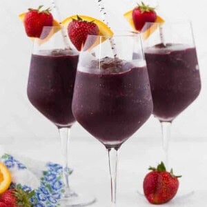 Three long stem wine glasses filled with frozen sangria, and topped with a fresh strawberry and orange slice. A paper straw is in each glass.
