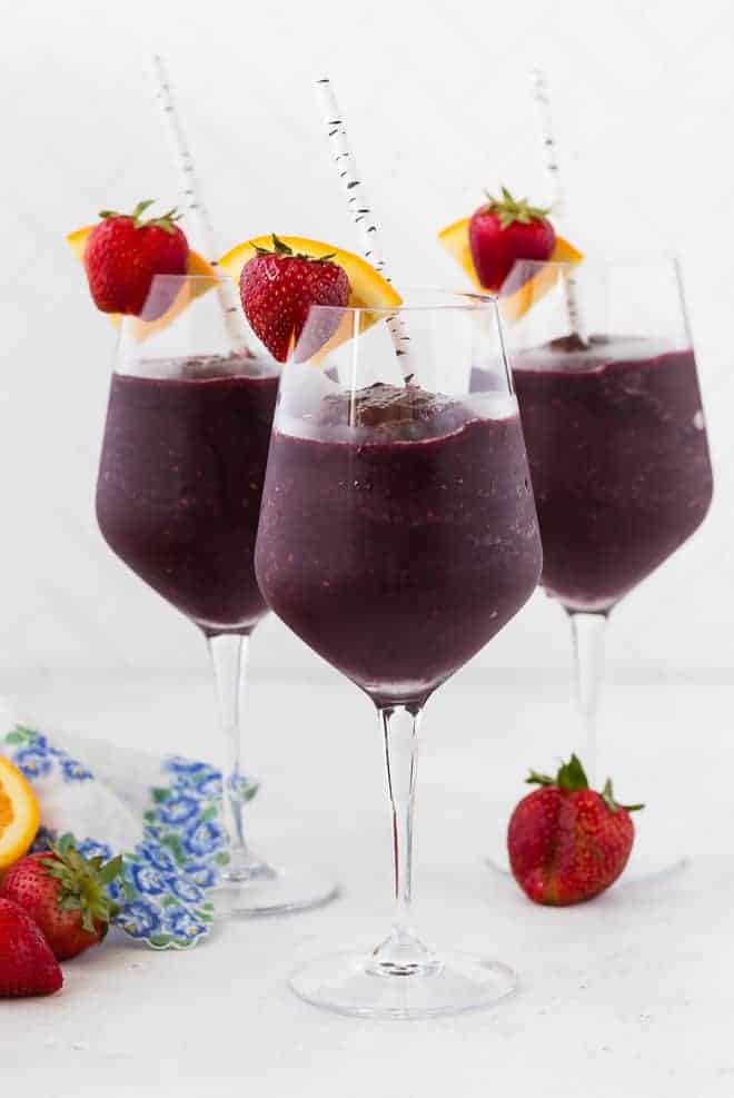 Three long stem wine glasses filled with frozen sangria, and topped with a fresh strawberry and orange slice. A paper straw is in each glass.