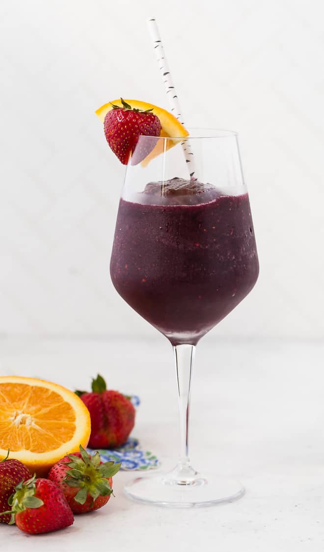 A long stem wine glass filled with a red wine sangria slushie, and topped with a fresh strawberry and orange slice. A paper straw is in the glass.