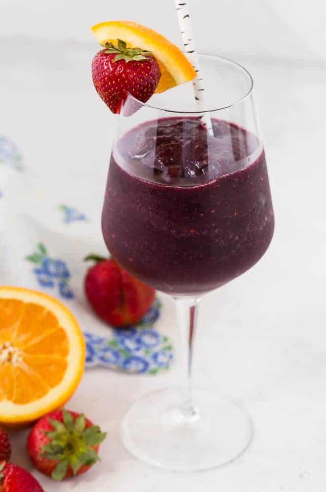 A long stem wine glass filled with a red wine sangria slushie, and topped with a fresh strawberry and orange slice. A paper straw is in each glass.