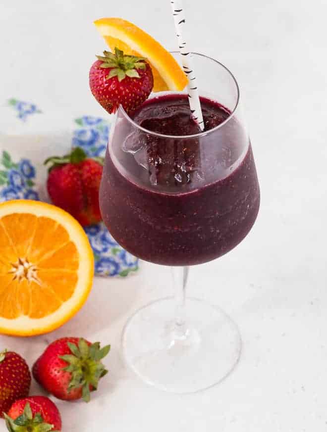 A wine glass filled with dark red wine slushie, surrounded by fresh oranges and strawberries.