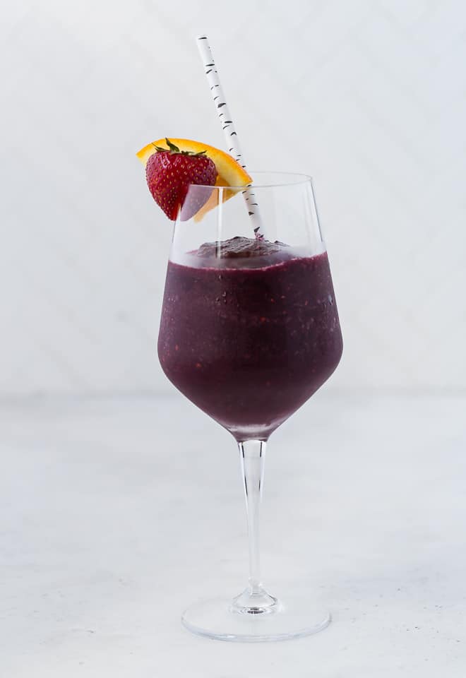 A long stem wine glass filled with a red wine sangria slushie, and topped with a fresh strawberry and orange slice. A paper straw is in the glass.